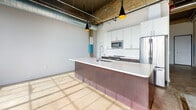 This 1-bedroom, 1-bathroom loft showcases a compelling blend of raw industrial character and contemporary design. The open-concept living and kitchen area is anchored by soaring exposed ceilings that reveal original concrete structure, ductwork, and piping — all hallmarks of authentic loft-style architecture. Polished and raw concrete floors run throughout, adding to the industrial aesthetic while remaining durable and low-maintenance.

The kitchen is a standout feature, centered around a generous island with a sleek white quartz countertop and a distinctive metallic base. White upper cabinetry pairs with dark lower cabinetry and a classic white subway tile backsplash, while stainless steel appliances — including a refrigerator, microwave, and range — complete the modern look. Stylish black pendant lights with gold interiors hang overhead, providing both task lighting and visual warmth.

Large industrial-grid windows flood the living area with natural light, casting dramatic shadow patterns across the floors while framing views of the surrounding cityscape. An original structural column with colorful painted accents adds a unique artistic touch to the bedroom.

The bathroom features white cabinetry, a vanity with sink, and clean, neutral finishes. Throughout the space, the interplay of raw materials and modern updates creates a dynamic, urban living environment.