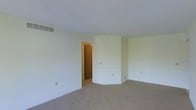 This single-floor space presents a straightforward and functional layout with a calm, neutral aesthetic throughout. The main living area is finished with wall-to-wall beige carpeting, lending a soft, cohesive feel underfoot. Walls are painted in a crisp off-white tone, contributing to a bright and open atmosphere, even in the absence of visible windows in this view.

A notable architectural detail is the angled wall section positioned toward the far end of the room, which adds subtle visual interest to what would otherwise be a simple rectangular layout. This angled feature creates a slight sense of dimensionality within the space. A doorway is visible, providing access to an adjoining room finished with warm wood-toned walls or paneling, hinting at additional living areas beyond.

Practical infrastructure is evident throughout, including a ceiling-mounted HVAC vent and multiple electrical outlets positioned at standard heights along the walls. The baseboard trim is clean and white, complementing the overall neutral palette. The space is currently vacant, allowing for a clear assessment of its structural elements and potential for flexible furnishing arrangements to suit a variety of functional needs.