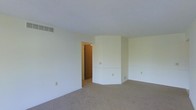 This single-floor space presents a straightforward and functional layout with a calm, neutral aesthetic throughout. The main living area is finished with wall-to-wall beige carpeting, lending a soft, cohesive feel underfoot. Walls are painted in a crisp off-white tone, contributing to a bright and open atmosphere, even in the absence of visible windows in this view.

A notable architectural detail is the angled wall section positioned toward the far end of the room, which adds subtle visual interest to what would otherwise be a simple rectangular layout. This angled feature creates a slight sense of dimensionality within the space. A doorway is visible, providing access to an adjoining room finished with warm wood-toned walls or paneling, hinting at additional living areas beyond.

Practical infrastructure is evident throughout, including a ceiling-mounted HVAC vent and multiple electrical outlets positioned at standard heights along the walls. The baseboard trim is clean and white, complementing the overall neutral palette. The space is currently vacant, allowing for a clear assessment of its structural elements and potential for flexible furnishing arrangements to suit a variety of functional needs.