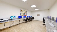 Located at 2401 6th St in Lake Charles, Louisiana, this computer center is a compact yet efficiently designed space that maximizes functionality within its single-floor layout. The room is finished with light hardwood-style flooring and crisp white walls, creating a bright and welcoming environment. Overhead, flush-mounted square panel lights provide consistent, even illumination throughout the space.

Long granite-topped wall-mounted desks line multiple walls of the room, offering ample workspace for numerous users simultaneously. Each workstation is equipped with a desktop computer, keyboard, and ergonomic mesh office chair, supporting a productive and comfortable user experience. A printer is also available, adding to the room's full-service amenities. A posted set of computer center rules is visible on the wall, indicating a structured and managed environment.

A vibrant abstract painting adds a touch of creativity and color to the otherwise clean and neutral interior. An additional granite-topped counter on the opposite wall provides extra surface space. The room is completed by a fire alarm panel and a security camera, ensuring safety and oversight. A glass-paned door leads to an adjacent area, contributing to the open and accessible feel of this well-organized, technology-focused space.