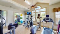 Located at 5799 Southland Dr in Mobile, Alabama, this fitness center is designed to provide a comprehensive workout environment in a welcoming, light-filled setting. The space is defined by its striking vaulted ceilings, which lend an open, spacious feel throughout the main exercise area. A prominent arched window crowns the front wall, flooding the room with natural light and offering pleasant outdoor views of the surrounding greenery, complemented by additional large multi-pane windows along the perimeter. A ceiling fan keeps the space comfortable during workouts.

The main workout area is well-stocked with a variety of cardio equipment, including treadmills, elliptical trainers, and stationary bikes, each equipped with individual monitor screens for entertainment. Wall-mounted televisions provide additional viewing options. A dedicated multi-station strength machine anchors one side of the room, while a colorful rack of stability balls, medicine balls, and resistance equipment adds versatility for functional training. An adjoining room, accessible through French doors with transom windows, houses a dumbbell rack for free weight training. The flooring throughout is a neutral, textured carpet suited for fitness use. A water cooler and posted fitness rules near the entrance reflect a well-maintained, community-oriented facility.