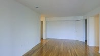This single-floor space presents a clean, minimalist aesthetic defined by warm-toned hardwood flooring and crisp white walls throughout. The main living area is generously proportioned, offering an open and flexible layout well-suited to a variety of furniture arrangements and uses. The hardwood floors display rich, natural grain patterns and provide a sense of warmth that contrasts with the bright, neutral wall surfaces.

The ceiling is smooth and white, featuring a centrally placed ventilation register, contributing to a tidy, uniform overhead finish. The walls are unadorned, allowing the space to feel airy and adaptable. Subtle architectural detailing includes a partial wall or column element that defines a portion of the room without fully enclosing it, adding mild structural character to the otherwise open plan.

Toward the rear of the space, a doorway provides access to an adjoining hallway or secondary room, suggesting additional functional areas beyond the main living zone. A door on the right side of the room hints at further storage or room options. Electrical outlets are positioned at baseboard level along the walls, offering practical placement for everyday use. Overall, the space conveys a straightforward, well-maintained character.
