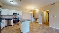 This single-floor residence is thoughtfully designed to offer a functional and comfortable living experience. The kitchen serves as the heart of the home, featuring white cabinetry, granite countertops, and a full suite of stainless steel appliances including a refrigerator, range, and microwave. A center island provides additional prep space and is complemented by a pendant light overhead. A built-in desk nook with open shelving adds a practical workspace seamlessly integrated into the kitchen area. Warm wood-look flooring runs throughout the kitchen and main living areas, creating a cohesive and easy-to-maintain surface. The living area is generously sized with neutral-toned walls and ceiling vents, offering a clean canvas for personalization. The bedrooms are finished with plush carpeting and feature vaulted ceilings, spacious walk-in closets with wire shelving systems, and direct access to the bathroom. A secondary bedroom benefits from a large window that fills the room with natural light and offers outdoor views. The bathroom is equipped with a soaking tub and shower combination, grab bars, wood-look flooring, and a vanity with storage. Overall, the space presents a clean, neutral aesthetic with practical features throughout.