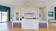 The Yardly Cross Creek Meadows sales center by Taylor Morrison is a thoughtfully designed single-floor community space located in Celina, TX. The open-concept layout is anchored by a sleek, minimalist reception desk positioned prominently at the entrance, with the Yardly branding boldly displayed on a crisp white feature wall — creating a polished and professional first impression.

Light-toned hardwood flooring runs throughout the space, complementing the bright white ceilings fitted with recessed lighting. A striking deep teal accent wall defines the lounge area, where leather seating arrangements and layered area rugs create a warm, inviting atmosphere for visitors. Large windows and floor-to-ceiling glass doors flood the interior with abundant natural light and offer direct views of the outdoor pool area and surrounding community.

A built-in workstation and refreshment nook with open shelving, a beverage cooler, and cabinetry provide practical amenities for guests. Near the entrance, a dedicated pet-friendly corner adds a welcoming community touch. The space also features additional seating areas and display walls showcasing community information and lifestyle imagery.

Overall, the center strikes a balance between contemporary design and functional comfort, offering visitors an informative and inviting environment to explore the Yardly Cross Creek Meadows community.