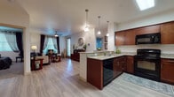 This single-floor residence offers a thoughtfully designed open-concept layout that seamlessly connects the kitchen and living areas. The kitchen is a standout feature, showcasing rich cherry wood cabinetry, granite countertops, a peninsula with pendant lighting overhead, and black appliances including a range, microwave, dishwasher, and refrigerator. Light wood-style flooring runs throughout the main living areas, enhancing the bright and airy atmosphere. The living room is generously sized, featuring a ceiling fan, multiple windows with draping curtains that invite natural light, and accent walls in a deep slate-blue hue that add character and warmth to the space. Two bedrooms provide comfortable retreats; the primary bedroom includes sliding glass doors with vertical blinds, plush carpeting, and a bold accent wall. Both bathrooms are finished with matching cherry wood vanities, granite countertops, large mirrors, and tub-shower combinations. A hallway connects the primary bathroom directly to the bedroom, offering a convenient layout. The unit also includes an entryway with additional storage and closet access. Overall, the space maintains a cohesive, modern aesthetic with warm wood tones, neutral walls, and practical finishes throughout.