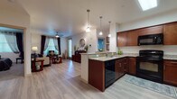 This single-floor residence offers a thoughtfully designed open-concept layout that seamlessly connects the kitchen and living areas. The kitchen is a standout feature, showcasing rich cherry wood cabinetry, granite countertops, a peninsula with pendant lighting overhead, and black appliances including a range, microwave, dishwasher, and refrigerator. Light wood-style flooring runs throughout the main living areas, enhancing the bright and airy atmosphere. The living room is generously sized, featuring a ceiling fan, multiple windows with draping curtains that invite natural light, and accent walls in a deep slate-blue hue that add character and warmth to the space. Two bedrooms provide comfortable retreats; the primary bedroom includes sliding glass doors with vertical blinds, plush carpeting, and a bold accent wall. Both bathrooms are finished with matching cherry wood vanities, granite countertops, large mirrors, and tub-shower combinations. A hallway connects the primary bathroom directly to the bedroom, offering a convenient layout. The unit also includes an entryway with additional storage and closet access. Overall, the space maintains a cohesive, modern aesthetic with warm wood tones, neutral walls, and practical finishes throughout.