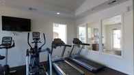 This single-floor fitness center is a dedicated workout space designed to support an active lifestyle. The room is thoughtfully laid out to accommodate a variety of cardio equipment, including multiple treadmills and an elliptical trainer, providing ample options for residents or users looking to maintain their fitness routines. A large wall-mounted mirror runs along one side of the room, creating a sense of expanded space while allowing users to monitor their form during exercise. The walls are finished in a clean, bright white, lending the space a fresh and energizing atmosphere. Recessed lighting in the tray ceiling supplements the abundant natural light that streams in through multiple windows, which frame views of the surrounding residential community. The windows not only brighten the interior but also provide pleasant outdoor sightlines, making workouts feel less confined. The flooring features a durable, dark-toned carpet suited for a fitness environment. A wall-mounted television adds convenience for entertainment during exercise sessions. Overall, the space is clean, functional, and well-lit, offering a comfortable and motivating environment for physical activity.