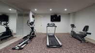The Regency Fitness Center is a dedicated workout facility designed to provide residents with a functional and comfortable exercise environment. The space is finished with neutral gray walls and a patterned carpet in muted tones accented with pink, creating a clean and practical aesthetic throughout. Recessed lighting illuminates the room evenly, contributing to a bright and energizing atmosphere.

The fitness center is thoughtfully equipped with a range of cardio and strength training equipment. Cardio options include a treadmill, an elliptical trainer, and a stationary bike, all positioned to allow ample space for use. A wall-mounted flat-screen television is installed above the cardio area, providing entertainment during workouts. For strength training, the facility features a multi-function cable machine, a weight rack loaded with plate weights, a barbell station, and a captain's chair for core exercises.

A full-length mirror spans one wall, enhancing the sense of space while allowing users to monitor their form. A workout reference chart is posted near the entrance for guidance. A sleek cylindrical waste receptacle is conveniently placed near the entry. The space is accessed through a secure door marked with fitness center signage, ensuring a private and dedicated environment for exercise.