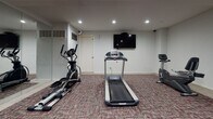 The Regency Fitness Center is a dedicated workout facility designed to provide residents with a functional and comfortable exercise environment. The space is finished with neutral gray walls and a patterned carpet in muted tones accented with pink, creating a clean and practical aesthetic throughout. Recessed lighting illuminates the room evenly, contributing to a bright and energizing atmosphere.

The fitness center is thoughtfully equipped with a range of cardio and strength training equipment. Cardio options include a treadmill, an elliptical trainer, and a stationary bike, all positioned to allow ample space for use. A wall-mounted flat-screen television is installed above the cardio area, providing entertainment during workouts. For strength training, the facility features a multi-function cable machine, a weight rack loaded with plate weights, a barbell station, and a captain's chair for core exercises.

A full-length mirror spans one wall, enhancing the sense of space while allowing users to monitor their form. A workout reference chart is posted near the entrance for guidance. A sleek cylindrical waste receptacle is conveniently placed near the entry. The space is accessed through a secure door marked with fitness center signage, ensuring a private and dedicated environment for exercise.