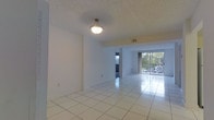 This single-floor space presents an open and airy layout highlighted by large white ceramic tile flooring that extends throughout the main living area, reflecting light and contributing to a clean, spacious feel. The white walls are smooth and neutral, providing a versatile backdrop adaptable to a variety of interior styles. A textured popcorn ceiling adds a classic touch overhead, complemented by a centrally hung flush-mount light fixture that provides ambient illumination to the room.

A standout feature of the living area is the full-width sliding glass door at the far end, which opens to an outdoor balcony. This element draws in abundant natural light and offers views of surrounding greenery, creating a pleasant connection between the interior and exterior. A HVAC vent is visible near the ceiling, indicating climate control throughout the space.

An adjoining room or hallway is accessible through an open doorway, suggesting a functional flow between living areas. The overall ambiance is bright, open, and well-lit, making it a practical and welcoming space well-suited for comfortable everyday living.
