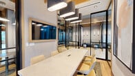 Located at 12004 Inspiration St in Reston, Virginia, this thoughtfully designed single-floor collaborative workspace offers a refined blend of modern aesthetics and functional versatility. Light oak hardwood flooring runs throughout, complemented by warm wood ceiling accents and exposed beams that lend the space an inviting, contemporary character.

The layout is organized around a central hallway corridor lined with sleek black-framed glass partitions, creating a sense of openness while defining distinct functional zones. A formal conference room features a large white marble-topped table surrounded by upholstered chairs, wall-mounted display screen, and striking oval pendant lighting overhead — ideal for productive meetings. Adjacent glass-enclosed lounge pods provide semi-private seating areas furnished with plaid bench seating, round ottomans, and slim wall sconces.

Deeper within the space, intimate booth seating alcoves set against large-format stone tile walls offer a quieter setting, each accented by sculptural column pendant lights. Additional flexible workspaces with task chairs and open tables are visible through the glass partitions. The corridor leads toward a set of glass entry doors that open to an exterior courtyard, flooding the hallway with natural light and creating a welcoming sense of arrival. The overall ambiance is polished, professional, and well-suited for collaborative or focused work.