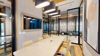 Located at 12004 Inspiration St in Reston, Virginia, this thoughtfully designed single-floor collaborative workspace offers a refined blend of modern aesthetics and functional versatility. Light oak hardwood flooring runs throughout, complemented by warm wood ceiling accents and exposed beams that lend the space an inviting, contemporary character.

The layout is organized around a central hallway corridor lined with sleek black-framed glass partitions, creating a sense of openness while defining distinct functional zones. A formal conference room features a large white marble-topped table surrounded by upholstered chairs, wall-mounted display screen, and striking oval pendant lighting overhead — ideal for productive meetings. Adjacent glass-enclosed lounge pods provide semi-private seating areas furnished with plaid bench seating, round ottomans, and slim wall sconces.

Deeper within the space, intimate booth seating alcoves set against large-format stone tile walls offer a quieter setting, each accented by sculptural column pendant lights. Additional flexible workspaces with task chairs and open tables are visible through the glass partitions. The corridor leads toward a set of glass entry doors that open to an exterior courtyard, flooding the hallway with natural light and creating a welcoming sense of arrival. The overall ambiance is polished, professional, and well-suited for collaborative or focused work.