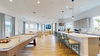 Located in Avondale, Arizona, this single-floor open-concept space seamlessly combines a fully equipped kitchen, dining area, and living room into one cohesive and inviting layout. Light wood-look flooring runs throughout, unifying the space and contributing to its airy, modern aesthetic.

The kitchen is a standout feature, showcasing soft gray shaker-style cabinetry, crisp white quartz countertops, a classic subway tile backsplash, and stainless steel appliances including a French door refrigerator. A large central island with bar seating provides an ideal gathering spot, while pendant lighting adds a stylish finishing touch. Multiple windows flood the kitchen with natural light and offer views of the surrounding landscape.

The living area is thoughtfully arranged with neutral-toned lounge seating, warm-toned accent décor, and a wall-mounted flat-screen TV atop a ribbed wood media console. A ceiling fan overhead ensures comfort, while large windows dressed in gray curtains with gold hardware frame views of palm trees and the neighborhood beyond.

Adding a recreational dimension, a full-size billiard table is centrally positioned in the shared space, making it equally suited for entertainment and relaxation. The overall ambiance is clean, bright, and welcoming — reflecting a contemporary Southwest design sensibility.