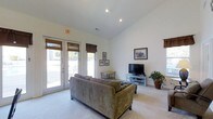 This single-floor space is a well-appointed common area designed for comfort and functionality. The room is characterized by soaring vaulted ceilings that lend an expansive, airy feel, complemented by recessed lighting throughout. Crisp white walls and neutral carpeting create a clean, inviting atmosphere.

The layout is thoughtfully organized into distinct zones. A lounge area features a plush, earth-toned sofa positioned to face a flat-screen television resting on a media console, making it an ideal spot for relaxation or casual gatherings. Adjacent to the lounge, a dining area accommodates a dark bistro-style table with matching chairs, suitable for small group meetings or social dining.

Large French doors with bamboo-style valances line the exterior wall, flooding the space with natural light and offering views of the outdoors. The doors also provide convenient direct access to an exterior patio or common outdoor area. Framed artwork decorates the walls, adding a touch of warmth and personality to the otherwise neutral palette.

A partial view of a kitchen or service area is visible along one wall, suggesting additional utility and convenience. The presence of exit signage indicates this is a shared or public-facing facility, making it well-suited as a clubhouse, leasing office lounge, or community gathering room.