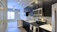This single-floor residence showcases a thoughtfully designed interior with a contemporary aesthetic. The kitchen is a standout feature, boasting rich dark cabinetry that contrasts beautifully with light countertops, creating a striking visual balance. A multicolored mosaic tile backsplash adds texture and character to the space, while stainless steel appliances — including a range, built-in microwave, and dishwasher — provide a polished, modern finish. Pendant lighting hangs elegantly above the counter, complementing the track lighting overhead and enhancing the kitchen's ambiance. The open-concept layout seamlessly connects the kitchen to the adjacent living area, which benefits from natural light streaming in through a window and is accented by warm hardwood-style flooring throughout. A ceiling fan in the living area adds both function and style. The overall design exudes a modern, urban feel, with clean lines, high-contrast finishes, and carefully chosen fixtures that come together to create a cohesive and inviting living environment.