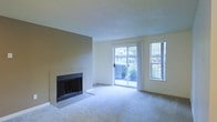This single-floor space centers around a well-appointed living room that combines functional design with a welcoming ambiance. A standout feature is the tile-surround fireplace set against a warm-toned accent wall, serving as a natural focal point for the room. The fireplace surround is finished in neutral tile, complementing the overall color palette of soft beiges and whites throughout the space.

The living area is carpeted in a light neutral tone, contributing to a warm and comfortable atmosphere underfoot. Natural light is generously introduced through a large sliding glass door, which provides direct views of surrounding greenery and outdoor scenery. An additional window positioned beside the sliding door further enhances brightness, ensuring the room remains well-lit throughout the day.

The sliding glass door offers convenient access to the outdoors while maintaining a visual connection with the natural environment beyond. The textured ceiling adds a subtle architectural detail consistent with the room's understated design. The overall layout is open and unobstructed, providing flexibility in furniture arrangement and making the space feel expansive and airy.