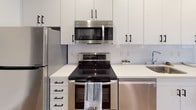 Located on the iconic Wilshire Boulevard in the heart of Los Angeles, this modern studio offers a thoughtfully designed open-plan layout that seamlessly integrates living, sleeping, dining, and cooking areas into one cohesive space. Warm hardwood-style flooring runs throughout, complementing the crisp white walls and clean-lined contemporary aesthetic.

The kitchen is well-appointed with full stainless steel appliances including a refrigerator, electric range, over-the-range microwave, and dishwasher, all paired with white shaker-style cabinetry accented by matte black hardware and a bright white tile backsplash. An in-unit washer is also conveniently included. The living area features large windows that invite natural light and offer views of the surrounding cityscape, with space for a sofa, dining table, and wall-mounted television on a wood media console. The sleeping area is comfortably appointed with a full-size bed and additional furnishings. The bathroom is finished in clean white tile and includes a combined shower and bathtub. Recessed lighting and modern fixtures complete the polished, urban feel of this well-located Los Angeles studio.