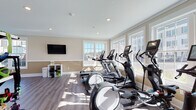 Located at 189 Paddlers Pt in Matamoras, PA, this single-floor fitness center is thoughtfully designed to support a comprehensive workout experience. The facility is divided into distinct zones, each tailored to a specific type of training, all unified by warm beige and white tones, durable hardwood-style flooring, and recessed lighting throughout.

The cardio area is generously sized and lined with large multi-pane windows that bathe the space in natural light and offer views of the surrounding community. This zone is equipped with multiple treadmills, elliptical machines, and a stair climber, along with a wall-mounted flat-screen TV for entertainment during workouts. Yoga mats and fitness accessories are neatly stored on open shelving nearby.

A dedicated strength training room provides ample space for free weight and resistance exercises. It features a full power rack with a weight bench, a cable machine, and additional free weights, complemented by its own wall-mounted television and a wall-mounted climate control unit. Crown molding and clean white trim add a polished finish to the room.

The layout, as seen in the overhead schematic, reveals a well-organized, multi-room floor plan that maximizes usability while maintaining an open, airy ambiance. Natural light pours in from multiple window orientations, creating an inviting and energizing environment ideal for daily fitness routines.