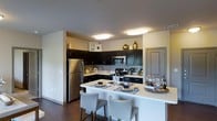 This single-floor space showcases a thoughtfully designed kitchen that serves as a central highlight. Dark espresso-toned cabinetry lines the walls in an L-shaped configuration, providing generous storage while creating a striking visual contrast against the light-colored walls and white countertops. The kitchen is equipped with a full suite of stainless steel appliances, including a French door refrigerator, an electric range, and a built-in microwave, offering both functionality and a cohesive, contemporary aesthetic. A mosaic tile backsplash adds a decorative accent behind the range, lending subtle texture to the space. The expansive white breakfast bar extends outward to create a casual dining area, accommodated by cushioned bar-height chairs in a neutral tone. Flush-mounted oval ceiling fixtures provide bright, even illumination throughout the kitchen. Dark hardwood-style flooring grounds the space and complements the cabinetry tones. The open layout promotes an airy, connected feel, making the kitchen both a practical workspace and an inviting area for entertaining or casual dining.