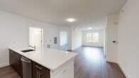This single-floor unit offers a clean, contemporary design highlighted by warm wood-tone flooring that flows consistently throughout the space. The open-concept layout connects the kitchen and living area seamlessly, creating a bright and airy atmosphere enhanced by natural light from well-placed windows. The kitchen is equipped with stainless steel appliances, including a gas range, over-the-range microwave, and refrigerator, complemented by dark wood cabinetry, white quartz countertops, and a stylish mosaic tile backsplash. A kitchen island with additional cabinet storage adds both functionality and a natural divide between the cooking and living spaces. The spacious living area provides ample room for a variety of furniture arrangements, with access to additional rooms via crisp white panel doors. The bedrooms are generously sized, each featuring wood-tone flooring, large windows with outdoor views, and closet access. Two full bathrooms serve the unit, each finished with wood-tone vanities, white quartz countertops, large mirrors, and tub-shower combinations clad in classic white subway tile with a decorative accent border. Light tile flooring in the bathrooms adds a clean contrast to the rest of the unit. The overall aesthetic is modern and cohesive, with a neutral palette throughout.