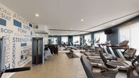 Located at 23641 Newhall Ave in Santa Clarita, California, this single-floor fitness center is a well-equipped facility designed to support a comprehensive workout experience. The space features a clean, modern aesthetic with white ceilings accented by recessed lighting, and bold teal accent walls that give the gym a vibrant, energetic atmosphere. A standout design element is the large typographic mural wall displaying fitness-themed words such as "Strength," "Athlete," "Endurance," and "Wellness," adding motivational character to the space. Full-length mirrors line one wall, visually expanding the room and allowing users to monitor their form during exercise. The cardio section is well-stocked with treadmills, elliptical trainers, and stationary bikes arranged in an orderly row along the windowed wall, where roller shades allow for natural light control. The strength training area features Matrix-brand cable machines, a weight bench, and a dumbbell rack, offering a solid range of resistance training options. Light tile flooring throughout keeps the space bright and easy to maintain. Glass panel partitions help define zones within the fitness center while maintaining an open, airy feel. Entry is through glass-paned doors, contributing to the space's welcoming and transparent design.