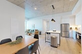 Located at 300 Mìwàte Pvt in Ottawa, this thoughtfully designed single-floor unit offers five bedrooms and four bathrooms within a modern, contemporary layout. The open-concept living, dining, and kitchen area forms the heart of the space, characterized by striking exposed concrete ceilings, light hardwood flooring, and an abundance of natural light streaming through floor-to-ceiling windows that frame city views. The kitchen is well-appointed with white cabinetry, stainless steel appliances including a refrigerator, stove, and dishwasher, under-cabinet lighting, and a central island with bar seating illuminated by sleek pendant lights. The dining area comfortably accommodates a full-sized table, flowing seamlessly into the living room where large glazed doors open to an outdoor balcony. Each of the five bedrooms is finished with clean white walls and hardwood flooring, offering built-in closet storage. The four bathrooms are uniformly designed with large-format stone-effect tiles, glass-enclosed walk-in showers, white vanities, and modern fixtures. Wide, bright hallways connect the private rooms, maintaining an organized and functional flow throughout the unit. The overall aesthetic is clean, modern, and urban.