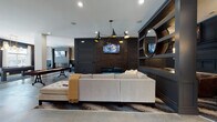 Located at 168 Headwater Circle in Mooresville, NC, this upscale community amenity space delivers a refined, modern aesthetic across a generously proportioned single-floor layout. Polished concrete floors unify the open-plan interior, reflecting light and lending an industrial-chic character throughout.

The centerpiece lounge area features a sleek linear fireplace set into a richly paneled dark accent wall, topped by a wall-mounted television. A plush sectional sofa and tufted ottoman anchor the seating arrangement, while a striking multi-arm brass and black chandelier adds dramatic overhead illumination. Built-in open shelving and a large round mirror frame the space with elegant detail.

Adjacent to the lounge, a stylish bar and kitchen area showcases a navy island with white quartz countertops, leather bar stools, and floor-to-ceiling open shelving fitted with pendant globe lighting. A white subway tile backsplash and warm wood ceiling accents provide contrast against the dark cabinetry.

The entertainment zone includes a billiards table and shuffleboard court, complemented by decorative lattice-panel accent walls. A casual dining area sits near large glass doors that open to the outdoors, filling the space with natural light.

A dedicated fitness center, accessible through full-glass double doors, is equipped with exercise machines and a mounted display screen, completing this well-appointed community destination.
