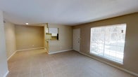 This single-floor space features an open-concept layout that seamlessly connects the living area to the kitchen. The living area is finished with light-toned ceramic tile flooring, contributing to a clean and airy feel. Neutral beige walls and white baseboards run throughout, creating a simple, cohesive aesthetic. A large window fitted with blinds allows abundant natural light to fill the space, offering a view of the exterior surroundings. The front entry door is centrally positioned along the main wall, providing straightforward access to the unit. The kitchen is partially separated from the living area by a raised pass-through counter, which adds a degree of definition between the two spaces while maintaining an open feel. White cabinetry lines the kitchen walls, and a sink is visible at the counter. The kitchen area transitions to carpeted flooring, distinguishing it subtly from the tiled living space. The ceiling features a textured finish throughout. Overall, the space presents a functional and practical layout with a neutral palette, making it adaptable to a variety of furnishing styles.