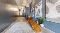 Located at 120 West Avenue in Kannapolis, NC, this thoughtfully designed lobby space makes a strong first impression through a carefully curated blend of modern aesthetics and inviting warmth. The entry is anchored by a striking set of black-framed glass double doors topped with a transom window, allowing abundant natural light to flood the space. Dramatic woven pendant chandeliers hang overhead, complementing the polished concrete floors that extend throughout.

The interior is finished in a sophisticated slate gray palette, providing a neutral canvas for standout decorative elements. A sculptural live-edge wood bench serves as a focal centerpiece, paired with large-scale abstract artwork displayed along the walls. Soft area rugs define seating zones and add texture underfoot.

Moving deeper into the space, circular ring pendant lights illuminate a connecting corridor, where a leather-upholstered bench with yellow accent pillows and lush potted greenery create a welcoming waiting area. A stacked stone accent wall adds an organic, tactile contrast to the otherwise sleek finishes.

The corridor leads to a pair of elevators flanked by vibrant large-format paintings, reinforcing the space's gallery-like character. Throughout, the design balances contemporary architecture with warm, natural materials, resulting in a polished yet approachable communal environment.