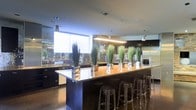 This sophisticated single-floor space showcases a seamlessly integrated modern design with a strong emphasis on clean lines and high-end finishes. The kitchen serves as the centerpiece, anchored by an expansive granite-topped island that offers generous counter space and is complemented by a row of clear acrylic bar stools, making it ideal for casual dining or entertaining. Dark, floor-to-ceiling cabinetry lines the perimeter walls, providing ample storage while reinforcing the space's sleek, contemporary aesthetic. A suite of stainless steel appliances adds a polished, professional-grade touch to the culinary area. Overhead, a combination of linear pendant lighting and individual pendant fixtures illuminates the space with precision, casting a warm glow across the reflective granite surfaces. A large picture window along one wall floods the kitchen with natural light and frames views of lush outdoor greenery, creating a striking contrast against the dark interior palette. Decorative tall grass arrangements displayed in glass vases along the island introduce organic texture and a touch of nature to the refined, monochromatic environment. The polished concrete-style flooring ties the entire space together, contributing to the overall sense of modern elegance.