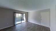 This single-floor space presents a clean, open layout highlighted by rich, dark hardwood-style flooring that contrasts neatly with the light gray walls and white baseboards throughout. The living area is generously proportioned, offering a versatile, open feel well-suited for a variety of furniture arrangements.

A prominent sliding glass door dominates one wall, providing direct access to a private balcony and drawing in an abundance of natural light. Sheer vertical blinds frame the door, allowing for adjustable privacy while maintaining the room's bright, airy atmosphere. The balcony beyond offers a partial outdoor view with surrounding greenery visible.

The ceiling features a classic textured finish, adding subtle visual depth to the space. Neutral gray walls create a versatile backdrop adaptable to various interior styles. A solid white entry door with a multi-point locking system is positioned on the adjacent wall, contributing to a sense of security and privacy. Electrical outlets and a cable or data port are conveniently placed along the walls. Overall, the space exudes a straightforward, functional ambiance with a fresh, move-in-ready aesthetic.