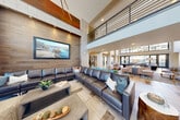 Located at 1607 17th St W in Billings, Montana, this expansive single-floor space spans a generously sized footprint designed for community gathering, collaboration, and everyday amenity use. The centerpiece of the space is a dramatic double-height lobby and lounge area, characterized by soaring ceilings, rich wood-paneled accent walls, and bold burnt-orange architectural details that draw the eye upward. Floor-to-ceiling windows flood the space with natural light, while a sleek open-grid shelving unit serves as a stylish room divider, separating intimate seating vignettes from a larger lounge furnished with an expansive dark leather sectional sofa. An upper-level mezzanine with metal railings overlooks the main living area, adding architectural depth and visual interest. A dedicated casual dining and kitchen area features a large central island with pendant lighting, dark navy cabinetry, and direct access to an outdoor terrace through oversized sliding glass doors, bringing in views of the surrounding exterior. Additional high-top communal tables provide flexible seating throughout. A private conference room, equipped with a large rectangular table, ergonomic seating, a projector, and glass partition walls, offers a professional setting for meetings. The overall ambiance blends contemporary design with warm, natural materials for an inviting and functional environment.