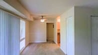 This single-floor residence offers a clean, functional layout designed for comfortable everyday living. The interior is finished in soft neutral tones throughout, creating a bright and welcoming ambiance that is further enhanced by natural light streaming in through windows along the walls. The main living area features light hardwood-style flooring that adds warmth and continuity to the space. A ceiling fan with a built-in light fixture anchors the room, providing both ventilation and overhead illumination. Architectural details include a subtle tray or beam detail in the ceiling, adding a touch of dimension to the otherwise smooth, open overhead space. A central hallway connects the main living area to the rest of the home, offering an organized flow between rooms. Just off the main space, a glimpse into the adjacent kitchen reveals warm wood cabinetry that brings a natural, inviting character to the cooking area. Clean white trim and door casings frame the openings throughout, contributing to a crisp, tidy aesthetic. A closeted room is also accessible, adding practical storage or additional functional space to the layout. Overall, the space presents a straightforward and well-maintained interior with a calm, neutral palette suited to a variety of living arrangements.