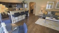 This single-floor residence showcases an inviting open-concept layout that effortlessly connects the main living areas. Rich, dark hardwood flooring flows throughout, anchoring the space with warmth and continuity. The kitchen is appointed with dark espresso cabinetry, granite countertops, and stainless steel appliances, including a built-in microwave. A raised breakfast bar with sleek chrome bar stools provides a casual dining option and serves as a natural divider between the kitchen and the living areas. The dining space accommodates a glass-top table and upholstered seating, perfect for everyday meals. The living room is comfortably proportioned, featuring a neutral sofa, a geometric-patterned area rug, and stylish accent furnishings that complement the warm beige wall tones. A distinctive arched doorway leads from the main living area into a secondary space, likely a home office or study, furnished with a desk and shelving. This architectural detail adds character and visual interest to the layout. Natural and ambient lighting work together to create a bright, welcoming atmosphere throughout. The overall design balances functionality with an understated elegance, making this a comfortable and well-appointed home.