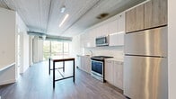 Located at 1590 Elmwood Ave in Evanston, IL, this modern one-bedroom, two-bathroom unit offers a thoughtfully designed open layout with contemporary finishes throughout. The combined kitchen and living area serves as the heart of the home, featuring an exposed concrete ceiling with industrial ductwork that lends an urban, loft-like character. The kitchen is equipped with stainless steel appliances, a gas range, a built-in microwave, and a combination of sleek white upper cabinets and light wood lower cabinets, complemented by a white tile backsplash. A butcher-block-topped island adds both function and warmth to the space. Large floor-to-ceiling windows flood the living area with natural light and offer treetop and city views, while a glass door provides access to an outdoor patio. The bedroom is a bright, well-proportioned retreat with wide windows overlooking the surrounding greenery and cityscape. The unit includes three closets for ample storage. Both bathrooms are finished with large-format tile flooring and wood-toned floating vanities; the primary bath features a dual-sink vanity with an oversized mirror, while the second bath offers a walk-in shower. Light wood-look flooring runs throughout the living spaces, tying the modern aesthetic together cohesively.