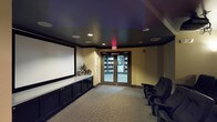 This private home theater is thoughtfully designed to deliver a true cinematic experience. The room features a striking dark ceiling with recessed lighting that creates an immersive, low-ambient atmosphere perfectly suited for screening films. A large projection screen dominates one wall, flanked by warm, neutral-toned walls that balance the room's dramatic aesthetic. Along one side, sleek dark cabinetry provides generous built-in storage with a clean, continuous countertop surface, ideal for media equipment or décor accents. The space is furnished with dedicated theater-style seating, offering a comfortable and authentic movie-viewing setup. Plush carpeting underfoot adds to the acoustic and comfort qualities of the room. A set of glass-paneled double doors serves as the main entry, offering a glimpse into an adjacent area and allowing controlled access while maintaining the room's enclosed, theater-like feel. Ceiling-mounted speakers are integrated seamlessly into the design, supporting a full audio experience. The overall ambiance is intimate and refined, combining functional design with an upscale aesthetic that makes this space an exceptional venue for private screenings and entertainment gatherings.