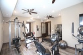 This single-level fitness center is designed to support a comprehensive workout experience within a thoughtfully arranged layout. The space is finished with light wood-style flooring and neutral beige walls, creating a clean, functional environment. Multiple ceiling fans are distributed throughout, ensuring consistent airflow during exercise sessions.

The facility is divided into distinct workout zones. One area houses an array of cardio equipment, including a treadmill, elliptical, recumbent bike, and upright cycle, all positioned near rows of windows fitted with horizontal blinds that allow natural light to filter in. A wall-mounted flat-screen television is also present in this zone for entertainment during workouts.

A dedicated strength training area features a multi-station cable machine, adjustable weight bench, flat bench, and a dumbbell rack with a stability ball, offering a range of resistance training options. A large wall mirror expands the visual space and allows users to monitor their form.

The entry area includes dark-stained wood doors, an exit sign, fire safety equipment, and a sanitation station, reflecting attention to safety and hygiene. An additional interior door leads to a separate room, adding further functionality to the overall facility.
