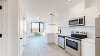 This thoughtfully designed single-floor unit showcases a clean, contemporary aesthetic throughout, characterized by crisp white walls, light-toned hardwood-style flooring, and an abundance of natural light. The open-concept layout seamlessly connects the kitchen and living area, creating a bright and airy atmosphere ideal for both daily living and entertaining.

The kitchen is a standout feature, outfitted with full stainless steel appliances — including a range, dishwasher, microwave, and refrigerator — paired with sleek white cabinetry that extends to the ceiling for ample storage. A peninsula with a white quartz countertop and a matte black gooseneck faucet anchors the space, complemented by a striking multi-bulb pendant light fixture. A large picture window in the adjacent living area frames city views and floods the space with natural light.

The bedroom offers a comfortable retreat with a generous walk-in closet and its own large window providing additional light and views. A wall-mounted PTAC unit ensures year-round climate control.

The bathroom is elegantly finished with white subway tile, a frameless glass shower panel, and coordinating matte black hardware and fixtures, lending a polished, modern feel. Throughout the unit, the consistent use of matte black accents, contemporary lighting, and high-quality finishes creates a cohesive and refined living environment.