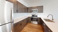This two-floor space is designed with a clean, modern aesthetic throughout. The staircase connecting both levels is finished with neutral carpet and bordered by crisp white walls, with a streamlined handrail guiding the way to the upper floor.

The kitchen is a standout feature, outfitted with dark espresso cabinetry, light quartz countertops, and a full suite of stainless steel appliances including a refrigerator, range, and microwave. Light wood-style flooring runs through the kitchen, adding warmth to the contemporary design.

The upper level reveals an open living area with light hardwood flooring and vaulted ceilings that lend a sense of spaciousness and volume. A large window with horizontal blinds allows natural light to fill the room while providing views of the surrounding exterior.

Bedrooms are carpeted for comfort, with neutral tones throughout. One bedroom features a ceiling fan, a window with blinds, and direct access to a generously sized walk-in closet equipped with wire shelving along multiple walls. Additional rooms on the upper level offer carpeted floors and clean white walls, providing flexible use.

A bathroom includes a walk-in shower with a white surround, wood-look flooring, and simple chrome fixtures, maintaining the consistent, understated style found throughout the space.