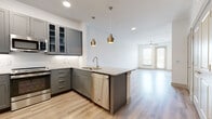 This thoughtfully designed single-floor residence showcases a contemporary aesthetic with a clean, neutral palette and quality finishes throughout. The open-concept kitchen is a standout feature, equipped with sleek gray shaker-style cabinetry — including glass-front upper cabinets — granite countertops, a white subway tile backsplash, and a full suite of stainless steel appliances including a range, microwave, refrigerator, and dishwasher. A peninsula with additional counter seating connects the kitchen to the living area, while a pair of warm brass pendant lights add an elegant touch overhead. Light wood-style plank flooring flows seamlessly through the main living spaces, complementing the bright white walls and recessed lighting that create an airy, well-lit atmosphere. The living area opens toward a glazed door with access to a balcony, bringing in generous natural light. The bedroom features plush carpeting, a ceiling fan, and a window with outdoor views, offering a comfortable and private retreat. The bathroom is appointed with dual granite-topped vanities set in matching gray cabinetry, large mirrors, and the same light wood flooring, maintaining a cohesive design language throughout the space. A hallway with a frosted glass door leads to additional areas of the residence, adding to the functional and well-organized layout of this modern home.