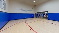 Located at 4300 Horizon North Pkwy in Dallas, TX, this professionally designed indoor basketball court is a standout athletic facility with a strong Dallas Mavericks theme throughout. The court features a classic light maple hardwood floor with bold red regulation markings, including a three-point arc and key, providing an authentic playing experience. Striking royal blue padded wall panels line the lower half of all walls, offering both protection and a vivid visual identity consistent with the Mavericks' team colors. A wall-mounted glass backboard and hoop anchor one end of the court, ideal for practice and competition alike. High ceilings flood the space with an open, airy atmosphere, complemented by recessed skylight-style ceiling fixtures that deliver bright, even illumination across the entire floor. Windows positioned near the upper walls allow additional natural light to filter in. Mavericks branding is prominently displayed, including retired jersey numbers for Nash (13), Nowitzki (41), Doncic (77), and Terry (31), along with the team logo and a large motivational quote attributed to Dick Vitale: "If you do the best you can, you have nothing to be ashamed of. A defeat is not a disgrace." A large action-photo mural and glass viewing panel complete the immersive fan experience, making this court as inspiring as it is functional.