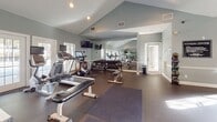 This single-story fitness center in Birmingham, Alabama offers a comprehensive workout environment designed to accommodate a variety of exercise routines. The space is characterized by vaulted ceilings that lend an open, airy feel, complemented by soft gray-green walls and white trim that create a clean, modern aesthetic throughout.

The facility is divided into distinct workout zones. The cardio area is anchored by multiple treadmills, elliptical trainers, and a stationary bike, each equipped with individual display screens. French doors and large windows along the perimeter flood this section with natural light and provide views of the surrounding outdoor area. Floor-to-ceiling mirrors along one wall visually expand the space while allowing users to monitor their form.

The strength training area houses a selection of cable machines, a dumbbell rack with a full range of free weights, flat benches, and a dedicated stretching mat. Recessed lighting and additional wall-mounted mirrors ensure the area remains bright and functional. A wall-mounted television adds convenience for entertainment during workouts.

The durable rubber flooring throughout provides both safety and comfort underfoot, while the overall layout allows for efficient traffic flow between equipment zones, making this fitness center a practical and well-appointed facility.