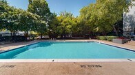 Located at Shady Point Square in Sterling, Virginia, this outdoor amenity space centers around a generously sized swimming pool with clear, blue water and a wide surrounding deck finished in a textured aggregate surface. The pool area is thoughtfully designed for safe recreational use, with designated entry points via metal handrails and clearly marked safety guidelines on the deck. A wrought iron perimeter fence encloses the entire area, providing both security and a defined boundary while maintaining an open, airy feel. Mature deciduous trees surround the space, offering natural shade and a lush green backdrop that enhances the outdoor atmosphere throughout the warmer months. A dedicated utility and restroom structure is situated adjacent to the pool, offering convenient amenities for guests. The gated entry to the pool area features ornamental ironwork consistent with the perimeter fencing, framed by flowering crape myrtle trees that add seasonal color. The residential buildings of the community are visible in the background, reinforcing the neighborhood setting. Overall, the space presents a well-maintained, community-oriented outdoor retreat designed for leisure and recreation under open skies.