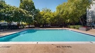 Located at Shady Point Square in Sterling, Virginia, this outdoor amenity space centers around a generously sized swimming pool with clear, blue water and a wide surrounding deck finished in a textured aggregate surface. The pool area is thoughtfully designed for safe recreational use, with designated entry points via metal handrails and clearly marked safety guidelines on the deck. A wrought iron perimeter fence encloses the entire area, providing both security and a defined boundary while maintaining an open, airy feel. Mature deciduous trees surround the space, offering natural shade and a lush green backdrop that enhances the outdoor atmosphere throughout the warmer months. A dedicated utility and restroom structure is situated adjacent to the pool, offering convenient amenities for guests. The gated entry to the pool area features ornamental ironwork consistent with the perimeter fencing, framed by flowering crape myrtle trees that add seasonal color. The residential buildings of the community are visible in the background, reinforcing the neighborhood setting. Overall, the space presents a well-maintained, community-oriented outdoor retreat designed for leisure and recreation under open skies.