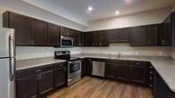 This single-floor space is thoughtfully designed with a clean, contemporary aesthetic that balances function and style. The kitchen serves as a standout feature, showcasing rich dark espresso-toned cabinetry that extends from base to upper wall units, providing generous storage throughout. Light granite-look countertops offer a striking contrast against the dark cabinetry, creating a visually appealing and practical workspace. The kitchen is fully equipped with a suite of stainless steel appliances, including an electric range, over-the-range microwave, dishwasher, and sink, making it well-suited for everyday cooking and entertaining. Warm hardwood-style flooring runs throughout, adding texture and a sense of continuity to the space. Recessed lighting in the ceiling provides bright, even illumination across the kitchen, enhancing both its functionality and ambiance. The neutral wall tones complement the overall color palette, contributing to a welcoming and modern atmosphere. The layout is efficient and open, allowing for comfortable movement and ease of use throughout the space.