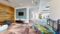 This contemporary fitness center is designed to offer a comprehensive workout experience within a single, well-appointed floor. The space is unified by a striking geometric-patterned carpet in vibrant shades of teal, green, yellow, and grey, which flows throughout the main corridors and exercise areas, lending a dynamic, energetic aesthetic to the environment.

The main corridor serves as the central spine of the facility, featuring a wall-mounted display set against a bold blue accent wall, warm wood-paneled walls, and an elevator for accessibility. This transitional space connects the various workout zones seamlessly.

The strength training area is generously proportioned, outfitted with a full rack of dumbbells, adjustable benches, cable machines, and weight stations. Full-wall mirrors expand the sense of space, while large windows allow abundant natural light and outdoor views to energize the room. Ceiling fans contribute to comfort during intense sessions.

The cardio zone houses a row of treadmills and elliptical machines, all positioned to take advantage of expansive exterior views through floor-to-ceiling windows. A dedicated functional training section with specialty equipment rounds out the gym floor.

A separate studio space stands out with its hardwood flooring, mirrored wall, and dramatic floor-to-ceiling glass doors that open to an outdoor terrace, flooding the room with natural light and fresh air — ideal for group fitness classes or individual training.