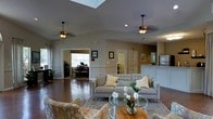 This single-story space offers an open and airy atmosphere, anchored by vaulted ceilings that lend a sense of generous vertical space. Two ceiling fans are positioned throughout the main living area, contributing both function and a classic aesthetic. Rich hardwood flooring extends across the space, grounding the warm, neutral color palette of the walls and furnishings.

The main living area is appointed with a contemporary gray sofa accented by patterned throw pillows, complemented by upholstered accent chairs in an earthy print. A glass-topped coffee table and a small round wooden side table serve as stylish, functional centerpieces. Recessed lighting and natural light from windows enhance the welcoming ambiance.

Adjacent to the living area, a defined room with a desk and seating is visible through an open doorway, suggesting a dedicated workspace or study. French doors nearby hint at an additional room, offering a sense of privacy and separation within the open floor plan.

Toward the rear of the space, a white counter-height divider separates what appears to be a kitchen or service area, keeping the layout connected yet organized. Overall, the space balances comfort with a clean, well-appointed design.