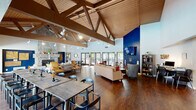 The Park San Dimas clubhouse is a generously sized community facility that serves as a welcoming social hub for residents. The centerpiece of the space is a dramatic great room distinguished by soaring vaulted ceilings with exposed wood beam trusses and warm wood paneling, all illuminated by track lighting. Rich hardwood flooring runs throughout, complementing the bold navy blue accent walls and crisp white walls. A floor-to-ceiling stacked stone fireplace anchors one end of the room, while a wall-mounted flat-screen TV occupies the opposite end. Multiple seating arrangements — including sofas, armchairs, and accent chairs — create inviting conversation areas, while a long communal dining table with chairs provides ample room for group gatherings. Banks of glass-paned doors and windows flood the interior with natural light and offer views of the outdoor pool area beyond. The clubhouse also includes a well-appointed kitchen with white shaker cabinetry, granite countertops, a subway tile backsplash, and stainless steel appliances. A dedicated laundry room provides multiple washers and dryers for resident use. A fitness area with motivational signage rounds out the amenities, making this clubhouse a comprehensive and vibrant community gathering space.