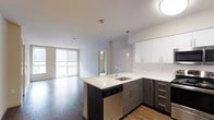 This contemporary apartment showcases an intelligently designed open floor plan that seamlessly integrates the kitchen and living spaces. The kitchen is a focal point, boasting dark wood cabinetry that contrasts beautifully with white upper cabinets and a light quartz countertop. A central island with a built-in sink serves as both a functional workspace and a subtle divider between the kitchen and living area. Stainless steel appliances, including a dishwasher, add a modern touch and practicality to the space. The living area features durable hardwood flooring that extends throughout, creating a cohesive look. Large windows flood the space with natural light, enhancing the sense of openness. Recessed lighting and pendant fixtures provide additional illumination options. A doorway visible in the background suggests access to private areas such as bedrooms or bathrooms. The neutral color palette and clean lines contribute to a fresh, uncluttered aesthetic that is both inviting and versatile for various decorating styles.