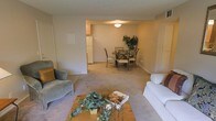 This single-floor residence offers an open and functional layout that seamlessly connects the living, dining, and kitchen areas. The living room is carpeted in a warm neutral tone and features a comfortable seating arrangement centered around a wooden coffee table adorned with decorative greenery. The space flows naturally into a dedicated dining area, where a round table with metal-framed chairs sits beneath a ceiling light fixture, accompanied by a potted plant that adds a touch of natural warmth. A partial wall separates the living and dining spaces without fully closing them off, maintaining an open and airy feel throughout. The kitchen is visible in the background, featuring light cabinetry and a white refrigerator. Walls throughout are painted in soft, neutral tones, complementing the carpeted floors and contributing to a cohesive, understated aesthetic. The overall ambiance is calm and practical, with a layout designed to maximize usability across each connected zone. Closet and storage access points are also visible, hinting at thoughtful built-in storage solutions throughout the home.